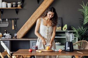 Charming curly-haired brunette girl in linen sundress prepares fruit smoothie in blender in kitchen. photo