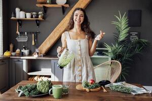 A happy curly-haired girl in a linen sundress is preparing a healthy breakfast. A young smiling woman holds a blender with a green smoothie. photo