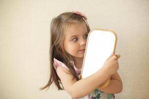 Young curious little girl holding mirror and looking at her reflection while playing alone at home photo