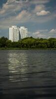 Two houses, multi-storey buildings, thick trees in front of them, and a large pond photo