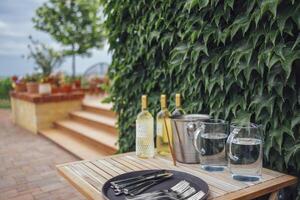 Three bottles of light wine, ice bucket, glass jugs of water, forks and spoons on tray on wooden table. Before garden party. photo