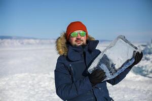Happy smiling man wearing winter blue jacket and red hat holding piece of transparent crushed ice photo