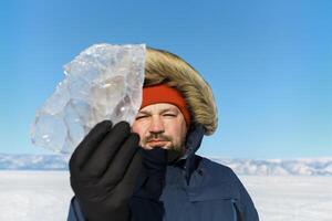 Happy smiling man wearing winter blue jacket and red hat holding piece of transparent crushed ice photo