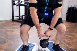 Strained man in ems suit standing on step platforms and doing squats with kettlebell. photo