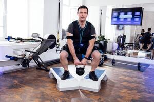 Strained man in ems suit standing on step platforms and doing squats with kettlebell. photo