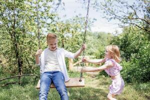 Schoolboy in jeans and white shirt is rocking his younger sister on swing. photo