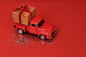 Red toy truck with craft gift box and red bow in its trunk. Miniature car with Christmas decoration on red bright background with sparkles and confetti. photo