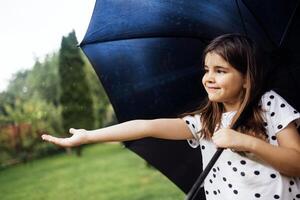 A little girl stands with a black umbrella in the rain. A cute baby stretches out her hand and catches raindrops in the park. photo