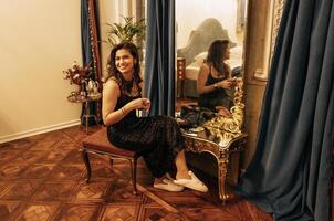 An attractive young woman in an evening dress and slippers is sitting in front of a dressing table and a large mirror. A smiling brunette girl is getting ready for a party. photo