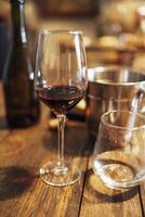 Close up of a still life of an elegant long-stemmed goblet with red wine and a glass of water on a textured wooden table. An ice cooler and a dark glass bottle in the background. Romantic atmosphere. photo