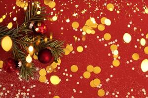 Branch of Christmas tree is decorated with red glass balls on red background. Bright backdrop with golden sparkles and round confetti. photo