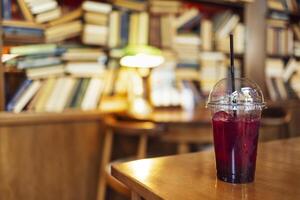A red lemon cocktail with ice cubes and a straw stands on a wooden table in the library. Large bookshelves, table, chair and lamp on the background at the cafe. Break while studying. photo