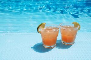 Two glasses with a tasty cocktail with ice cubes. Delicious fresh drink with lime slices by the pool. A sunny day and the reflection of rays in the blue water in the background. photo