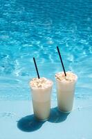 Two glasses with a tasty chilled cocktail. Milkshake with whipped cream. Delicious fresh drink by the pool. A sunny day and the reflection of rays in the blue water in the background. photo
