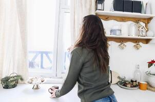 Happy woman looking out of kitchen window and drinking hot tea, enjoying New Year holidays at home. photo