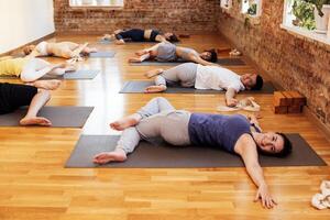 Morning yoga class with an instructor at the fitness club. A group of young multiracial people lie on mats and do Jathara Parivartanasana. Guys and girls do stretching or pilates. photo