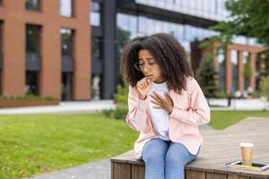 Young woman coughing while sitting outside urban building. Appears unwell and holding chest, suggesting possible respiratory issue or cold. Outdoor park setting with trees and modern architecture photo