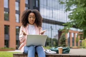 Frustrated student using laptop outdoors on campus. Displaying confusion or technical difficulties while studying. Urban background with modern buildings and green spaces. photo