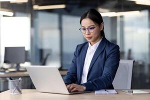 Businesswoman in formal attire working on laptop in modern office. Focused and diligent, she utilizes technology to accomplish tasks efficiently. Office environment reflects modern corporate setting. photo