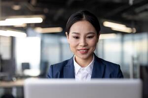 Smiling businesswoman working on laptop in modern office environment. Professional woman in formal attire focused on computer screen, representing productivity and corporate culture in workplace. photo