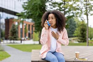 Young woman with asthma using inhaler for relief while sitting on bench in park. She looks anxious and is wearing casual clothes. Green trees and modern buildings in background. photo