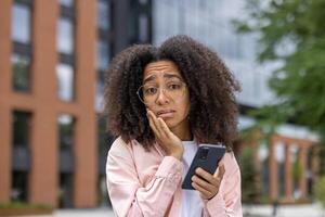 Young woman with glasses displaying expressions of confusion and worry while holding smartphone in urban setting. Emotion of uncertainty captured outdoors. Background features modern buildings photo