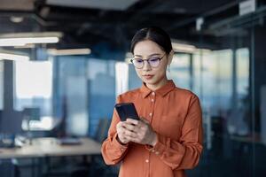 Asian woman using phone, serious thoughtful business woman inside office typing text message and browsing social media, female programmer software developer testing app online. photo