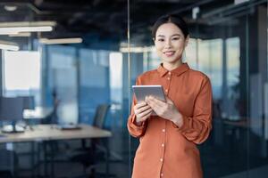 Portrait of a successful Asian female programmer with a tablet computer in her hands, an employee of a development company is smiling and looking at the camera near the window. photo
