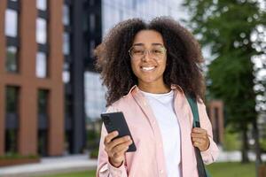 Happy woman student with glasses holding smartphone outdoors in urban setting. Youthful, cheerful expression showcasing relaxation and digital connectivity near modern building. Concept of education, technology, and casual lifestyle. photo