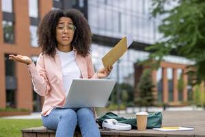Confused student holding envelope while using laptop. Outdoor workspace in urban setting. Person expressing confusion or misunderstanding. Concept of remote study and modern education methods. photo