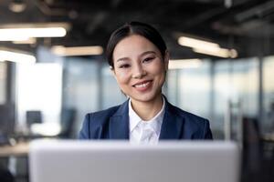 Asian businesswoman in formal attire working on a laptop in a modern office setting. She is smiling confidently, embodying professionalism and positivity. photo