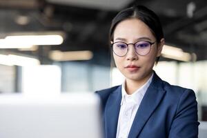Professional asian businesswoman wearing glasses and formal attire, concentrating on her work while using a laptop in a contemporary office setting. The image depicts focus, productivity. photo