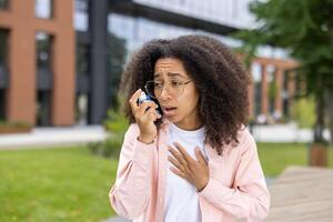 African American woman with using asthma inhaler outdoors. Young woman looking concerned or anxious while using inhaler to manage asthma symptoms. Concept of asthma, health, and respiratory issues photo