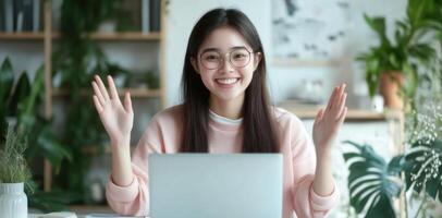 Asian woman sitting at a white desk using a laptop and waving her hand. photo