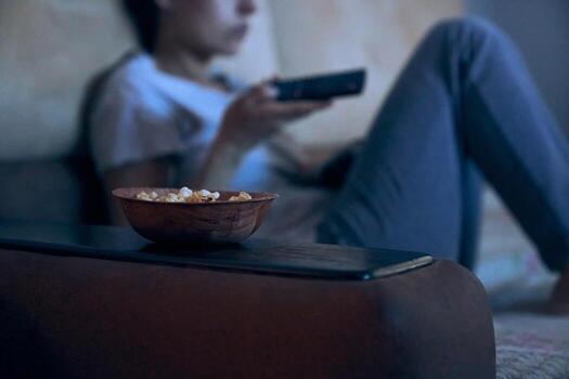 Cropped view of blurred woman sitting on sofa with remote control watching TV. Selective focus on plate with popcorn. Concept of insomnia, addiction, depression, problems. Background with copy space photo