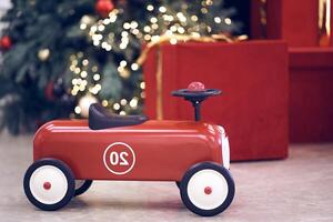 View of red children's retro car on the background of gifts in red boxes with golden bows, Christmas tree decorated garland, string light, red and blue balls. Background, copy space, bokeh photo