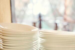A set of white plates on the table on a light blurred background of the window. Stacks of white dishes on hand at the hotel. photo