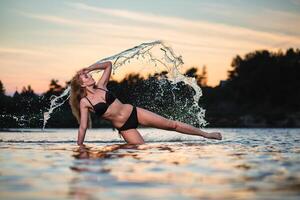 Young slender girl in a black swimsuit posing in the water at sunset at the background of water flying in the air photo