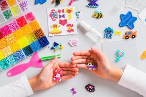 Children's hands holding toys made of colorful aqua beads and craft kit on table top view photo