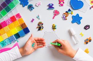 A girl makes a pattern of aqua beads using tweezers on a pegboard top view photo