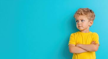 A young boy with a serious expression crosses his arms against a vibrant blue background, showcasing emotions and childhood innocence. photo