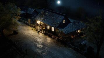 An aerial view of a small wooden cottage at night with a full moon and stars in the sky. The cottage is illuminated with warm light from the windows and a porch light. A path leads to the cottage through a forest. photo