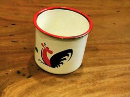 enamel cup with classic rooster painting pattern isolated on wooden table. vintage stuff. retro design for coffee shop decoration photo