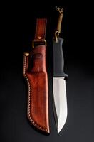 A modern hunting knife and a leather case for him on a dark background. Melee weapons for hunting and self-defense. The instrument of crime. photo