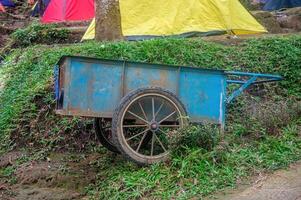 A vintage blue wheelbarrow sits abandoned in a grassy campsite surrounded by colorful tents. photo