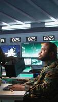 Vertical Military agent working in control room, analyzing aerial reconnaissance data and talking with colleague. Intelligence corps agent checking screens, discussing with coworker, camera B video