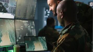 Military soldier in mission control center, overseeing national safety operations using notebook next to colleague. Team of army specialists examining data in monitoring room command center, camera A video