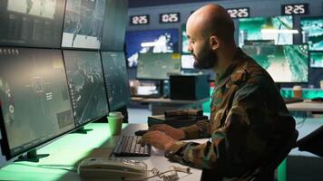 Military soldier in mission control center overseeing national safety operations with devices, close up. Middle Eastern army specialist examining data in monitoring room command center, camera B video