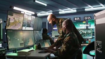 Military soldier in mission control center, overseeing national safety operations using notebook next to colleague. Team of army specialists examining data in monitoring room command center, camera B video