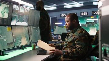Military soldier in mission control center overseeing national safety operations, reading folder files. African american army specialist checking data in monitoring room command center, camera B video